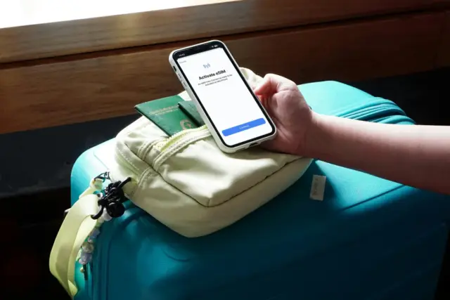 Traveler activating an eSIM on a smartphone screen while holding the phone beside a suitcase and fanny pack with passport and travel documents, upon arrival to Japan.