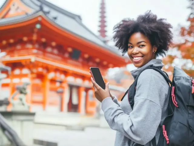 Cheerful young woman holding a smartphone in front of a Japanese Temple.