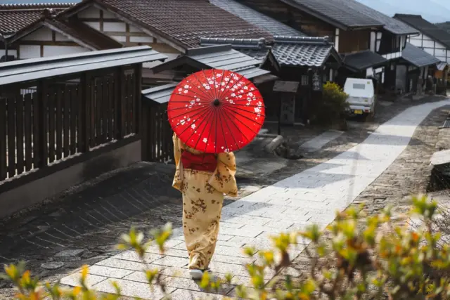 A woman wearing a traditional kimono, holding a paper umbrella while walking through a quiet street lined with traditional Japanese buildings.