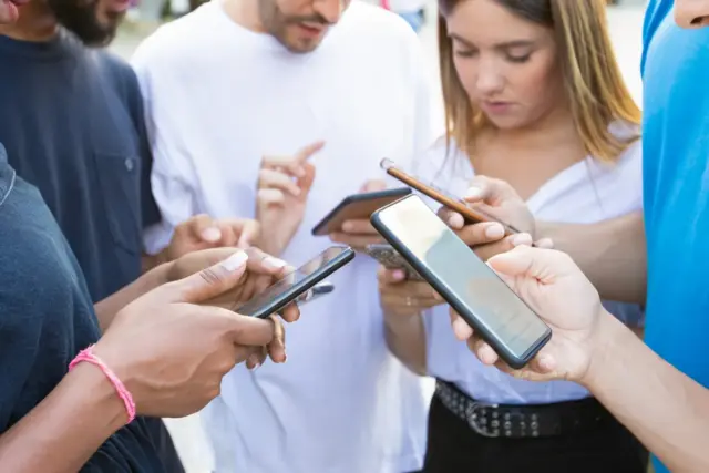 Group of five friends traveling in Japan, standing close together outdoors, looking down at their smartphones.