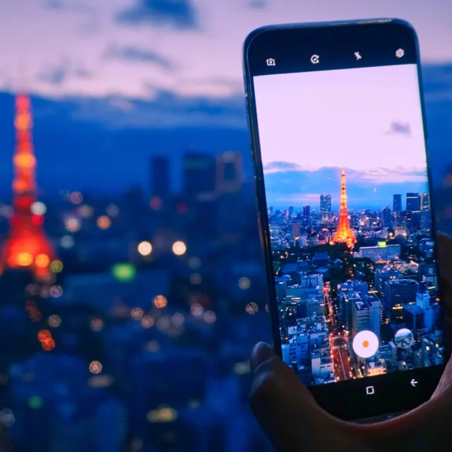 A hand holding a smartphone, recording the Tokyo skyline with Tokyo Tower at night.