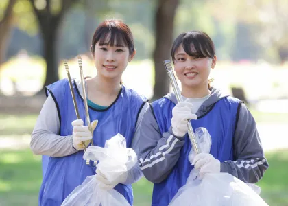 Two Japanese women holding tongs and trashbags, picking up trash in a park.