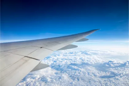 Aerial view from an airplane window showing the white wing extending over a vast, thick layer of white clouds against a clear blue sky.