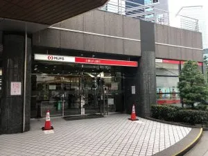 Exterior of a Mitsubishi UFJ (MUFG) bank branch in Japan, featuring the red and white corporate logo above a glass entrance with safety cones.