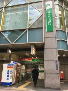 Entrance to a Japan Post Bank (Yucho Bank) branch, identified by its iconic green signage and a red mailbox located outside a modern office building.