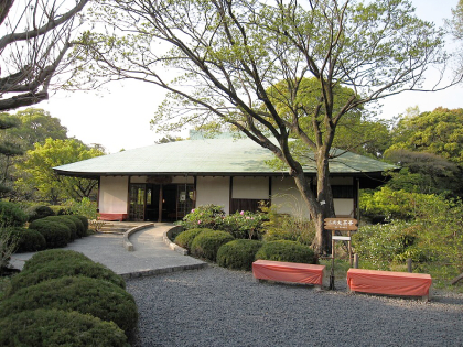 Traditional Japanese teahouse with green hipped roof surrounded by gravel courtyard and well-maintenance bushes