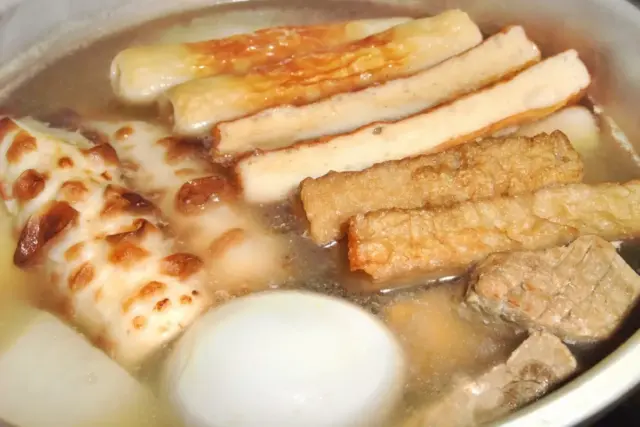 A steaming bowl of traditional Japanese oden featuring boiled egg, chikuwa fishcakes, and sliced fried fish cakes in a savory broth