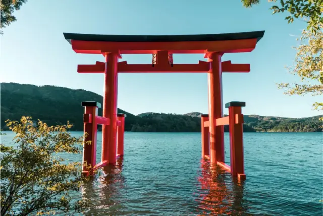 The vibrant red Heiwa no Torii gate standing in the calm waters of Lake Ashi with lush green hills in the background